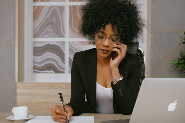 woman sitting at desk wearing a blazer and writing something down while on the phone