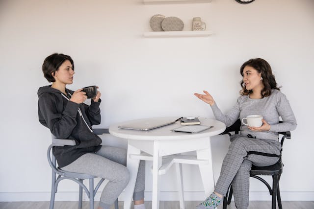 two people sitting at a white table having a conversation holdig coffee cups